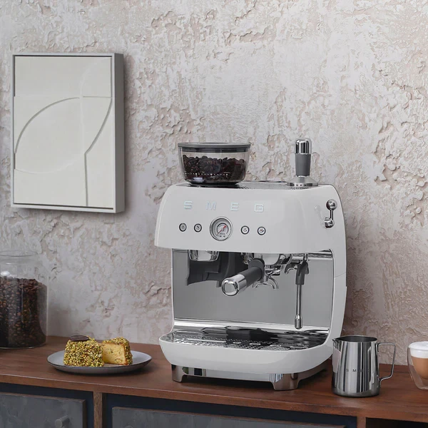 Aparat za kafu SMEG EGF03WHEU u bijeloj boji na drvenoj površini uz kolače i šoljicu kafe